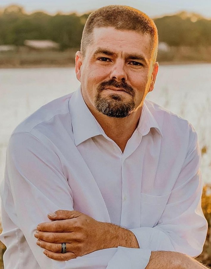 Portrait of Eric Halstead in a white shirt, arms folded, with a lake behind him at sunset.